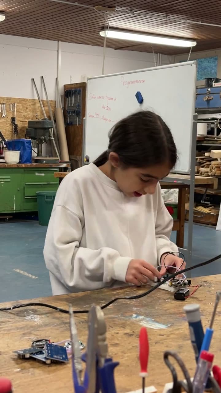 A child wires a small circuit board at a workbench, a whiteboard in the background